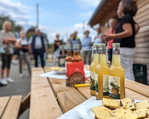 Holztisch mit Säften und belegten Broten. Personen stehen im Hintergrund.