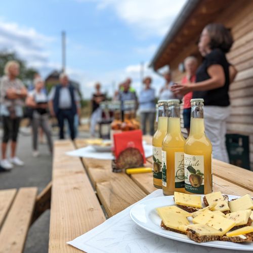 Holztisch mit Säften und belegten Broten. Personen stehen im Hintergrund.